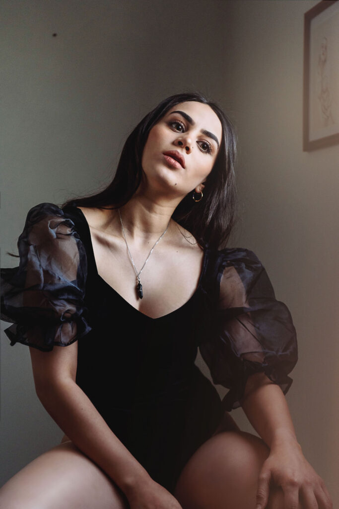 Valeria Cisneros poses elegantly in a room with soft lighting wearing a black dress with sheer puff sleeves a necklace and earrings.
